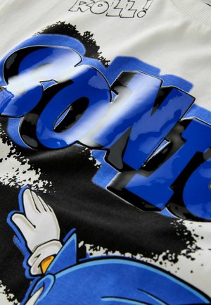 Weißes Baumwoll-T-Shirt mit fettem blauen "SONIC"-Text, glänzender Oberfläche und einer Grafik eines blauen Charakters, der winkt, vor einem schwarzen Spritzdesign.