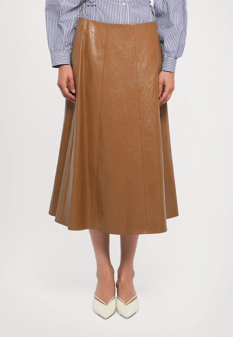 Falda midi de cuero marrón con una textura suave, que presenta costuras verticales y una silueta acampanada, combinada con tacones blancos de punta afilada.