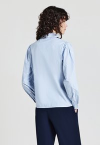 Person with curly dark hair wearing a light blue striped long-sleeve shirt and dark pants, standing with hands in pockets facing sideways.