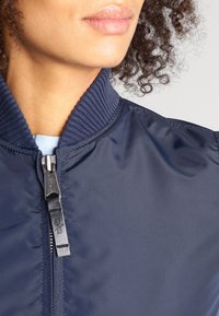 Granatowa kurtka bombowca z prążkowanym kołnierzem, zamkiem błyskawicznym, gładkim, błyszczącym materiałem, zdobiona subtelnymi przeszyciami i o luźnym kroju.