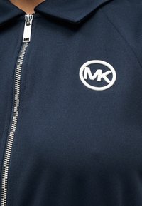 Μπλε μπουφάν με φερμουάρ και γιακά, με ασημένιο φερμουάρ και κεντημένο λευκό κυκλικό λογότυπο "MK" στα αριστερά. Λεία υφή.