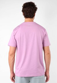 T-shirt lilas à manches courtes avec un col rond, présentant une petite patte de logo sur l'épaule gauche. Texture lisse et design décontracté.