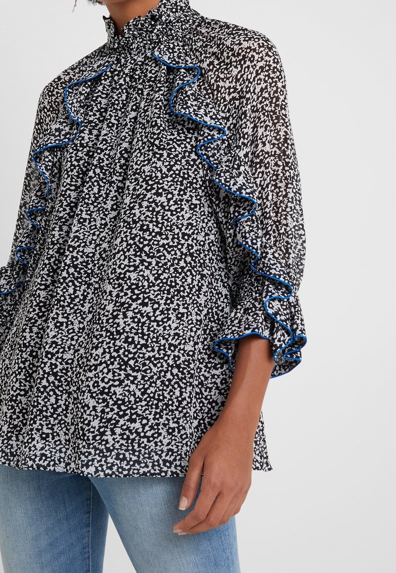 Blouse tachetée noir et blanc avec des volants bordés de bleu sur les épaules et les manches, portée avec un jean bleu.