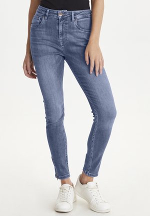 Niebieskie jeansy slim fit z suwakiem przy kostkach, noszone z białymi trampkami i czarnym topem, pokazane od talii do stóp.