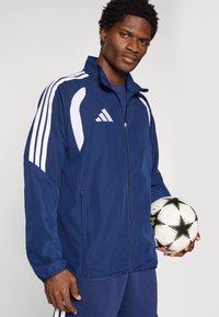 Μπλε ναυτικό μπουφάν με φερμουάρ, με λευκές λεπτομέρειες, που διαθέτει τρεις ρίγες στα μανίκια και τσέπη με φερμουάρ. Το μοντέλο κρατάει μια ασπρόμαυρη ποδοσφαιρική μπάλα.