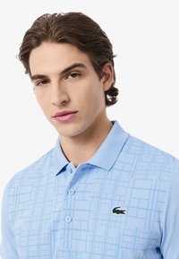 Polo bleu clair avec un motif géométrique, deux boutons et un logo crocodile vert sur la poitrine gauche. Matière en coton lisse.