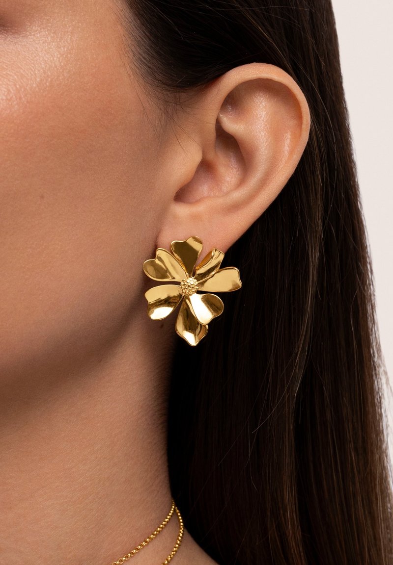 Boucle d'oreille dorée en forme de fleur avec cinq pétales portée à l'oreille d'une personne aux cheveux lisses et foncés et à la peau lisse.