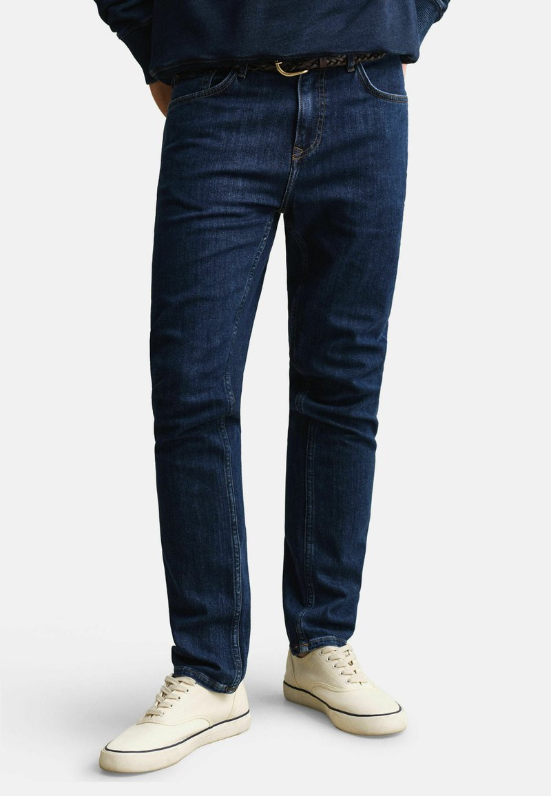 Hombre con vaqueros ajustados azul oscuro, un cinturón trenzado marrón y zapatillas deportivas blancas con cordones, de pie frente a un fondo blanco.