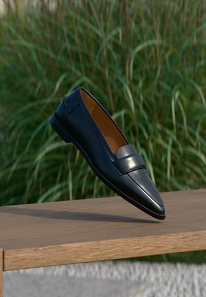Men’s black leather loafer with strap detail balancing on the edge of a wooden bench against blurred green grass background.