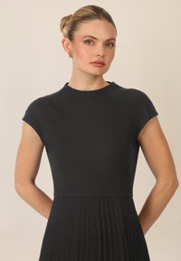 Vestido negro de manga corta con parte superior acanalada y falda plisada. Textura suave y cuerpo ajustado, con escote redondo. Pendientes dorados.