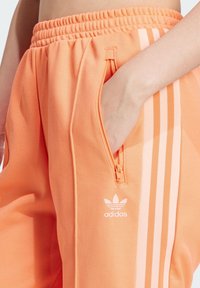 Coral orange Adidas pants with a textured finish, featuring a zippered pocket and three white stripes along the side.