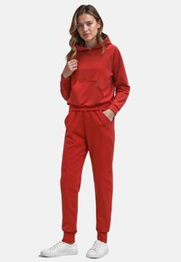 Felpa rossa con tasca a marsupio e pantaloni coordinati, realizzati in tessuto morbido. Presenta polsini e vita elastici, abbinati a sneaker bianche.
