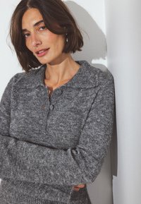 Sivý pletený sveter s golierom a dvoma gombíkmi. Mäkkučká, textúrovaná látka. Model stojí proti bielej stene s prekríženými rukami.