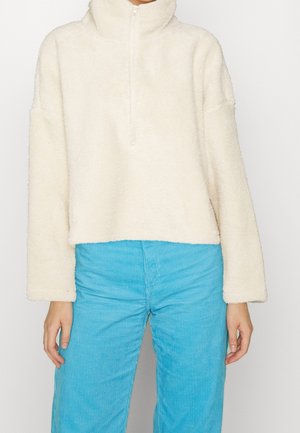 Person iført en cremefarvet fleece-pullover med halvlukning og lyseblå fløjlsbukser, stående foran en ensfarvet baggrund.