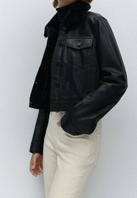 Chaqueta de cuero negra cortada con un cuello de piel, con botones frontales y un bolsillo en el pecho, combinada con pantalones beige claros.
