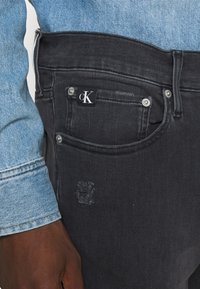 Jeans de mezclilla negros con un pequeño parche de logo visible, bolsillos frontales y detalles deshilachados. Combinados con una chaqueta de mezclilla azul claro.