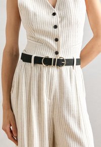 Ceinture en cuir noire avec double boucle en or, portée sur une tenue en tissu à rayures crème avec un décolleté en V et une patte de boutons.