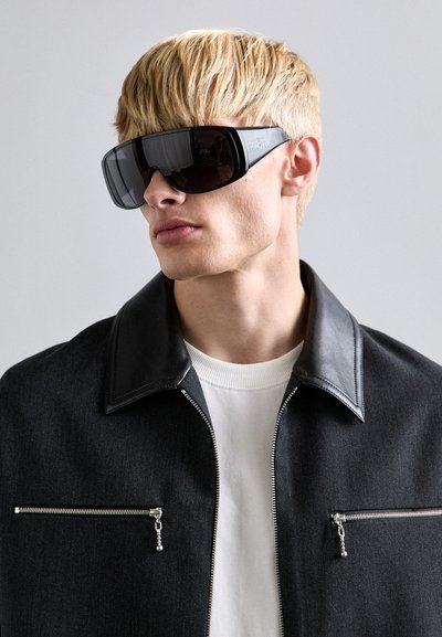 Óculos de sol grandes e pretos com um design elegante. O modelo usa uma jaqueta preta com colarinho de couro e um suéter branco por baixo.