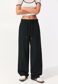Vrouw in zwarte wijde broek met trekkoord, wit cropped t-shirt met zwarte rand en witte sneakers, staand met gekruiste armen.