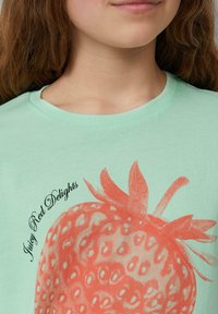 Jasnozielona koszulka z grafiką koralowej truskawki i czarnym napisem "Soczyste Czerwone Przyjemności". Okrągły dekolt i miękki bawełniany materiał.