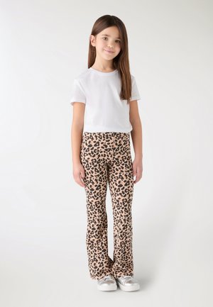 Dziewczyna stoi, ubrana w białą koszulkę, spodnie w panterkę i srebrne trampki na jednolitym jasnym tle.