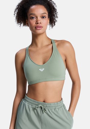 Młoda kobieta o kręconych włosach, ubrana w miętowozielony sportowy stanik i pasujące prążkowane spodnie z troczkiem na białym tle.