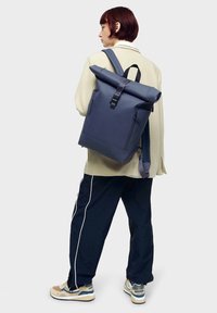 Jūras zila rullējamā mugursoma ar matētu apdari, regulējamiem siksnām un priekšējo sprādzi. Ietver sānu kabatu uzglabāšanai.