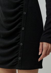 Schwarzes Kleid mit gerafftem Stoffdetail auf einer Seite, sechs schwarzen Knöpfen, die vertikal entlang der Naht angeordnet sind, und langen Ärmeln. Weicher Stoff.
