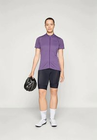 Žena v cyklistickom oblečení drží čiernu prilbu, má oblečené fialové tričko s krátkym rukávom, čierne šortky, biele ponožky a biele topánky.