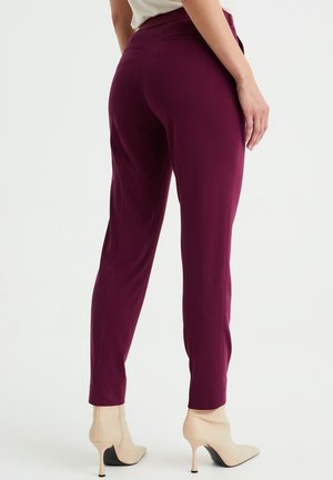 Femme portant un pantalon moulant violet foncé et des bottines beige à talons hauts, vue de dos sur un fond uni.