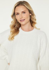 Pulover alb din tricot cu model de cablu, cu guler rotund și mâneci lungi. Materialul pare texturat și moale, având un model reliefat.