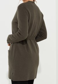 Cardigan long en maille vert olive avec une taille ceinturée, des poches latérales et une coupe décontractée. Texture douce et manches longues.