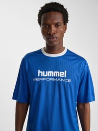 Unge sort mand med kort krøllet hår bærer en blå "hummel PERFORMANCE" sportst-shirt med hvid krave mod en ensfarvet hvid baggrund.