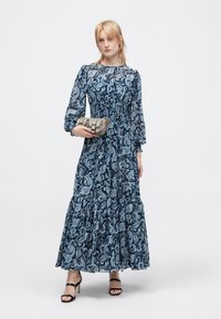 Vestido longo em paisley azul escuro com mangas longas e franzidas e saia em camadas. Acessorizado com uma pequena bolsa prateada e sapatos de salto pretos com tiras.