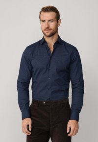 Man in een strak passend donkerblauw gestreept overhemd en donkerbruine broek, staande tegen een effen lichte achtergrond.