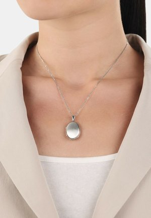 Silbermedaillon-Halskette mit einem polierten ovalen Anhänger an einer zarten Kette, getragen zu einem hellen Oberteil und Blazer.
