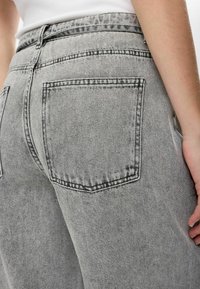 Gros plan d'une personne portant un jean gris taille haute et un haut blanc, montrant la poche arrière et le bras latéral avec la main reposant.