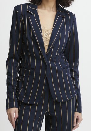 Frau trägt einen marineblauen Anzug mit dünnen beigen vertikalen Streifen und einem beigen Spitzenoberteil darunter, steht vor einem einfarbigen Hintergrund.