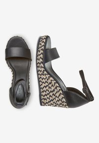 Sandalias de cuña negras con un diseño de patrón trenzado, que cuentan con una correa de cuero suave y una suela texturizada. Correa de tobillo ajustable para un mejor ajuste.