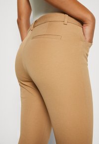 Pantalons stretch beiges avec une texture lisse, dotés d'une taille mi-haute, de deux poches arrière et d'une silhouette ajustée.