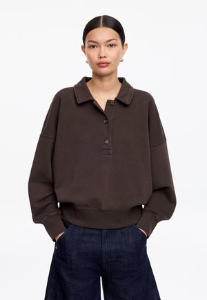 Femme aux cheveux bruns foncés, raie au milieu basse, portant un pull à col boutonné marron foncé et un jean bleu à jambes larges, les mains dans les poches.