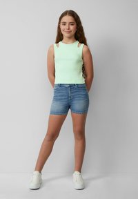 Jasnozielony top bez rękawów z wycięciami na ramionach, w zestawieniu z niebieskimi dżinsowymi szortami i białymi trampkami, stojący na neutralnym tle.
