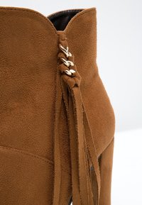 Hnědé semišové kotníkové boty s akcenty zlatého řetízku a semišovými střapci po straně, s vysokým podpatkem a zvlněným horním okrajem.