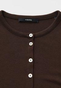 Brauner Strickcardigan mit rundem Ausschnitt und vier weißen Knöpfen auf der Vorderseite, mit einem schwarzen Etikett, das innen am Kragen eingenäht ist.