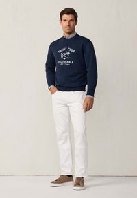 Felpa navy con testo bianco e grafiche nautiche, abbinata a pantaloni bianchi e sneakers in suede marrone. Camicia a fantasia visibile sotto.