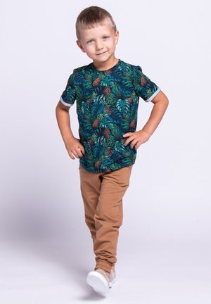 Koszula z krótkim rękawem zdobiona zielonym tropikalnym liściem i brązowym wzorem ananasa, zestawiona z beżowymi spodniami i jasnoszarymi tenisówkami.