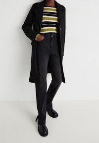 Abrigo de lana negro sobre un suéter de canalé a rayas en verde, negro y blanco. Jeans de mezclilla negros con lazos laterales. Botas de plataforma negras.