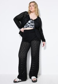 Zwarte gehaakte cardigan met open breipatroon, gecombineerd met een bijpassende zwarte cropped top en losse broek, gedragen met witte sandalen.