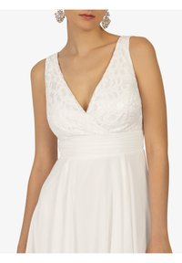 Robe blanche avec un col en V et un corsage en dentelle élaborée. Jupe lisse et fluide avec une taille cintrée. Boucles d'oreilles pendantes argentées mettent en valeur le look.