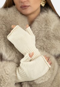 Beżowe bezpalcowe rękawiczki o prążkowanej fakturze i otworze na kciuk, wykonane z miękkiego dzianinowego materiału, trzymane na tle puszystego futra.